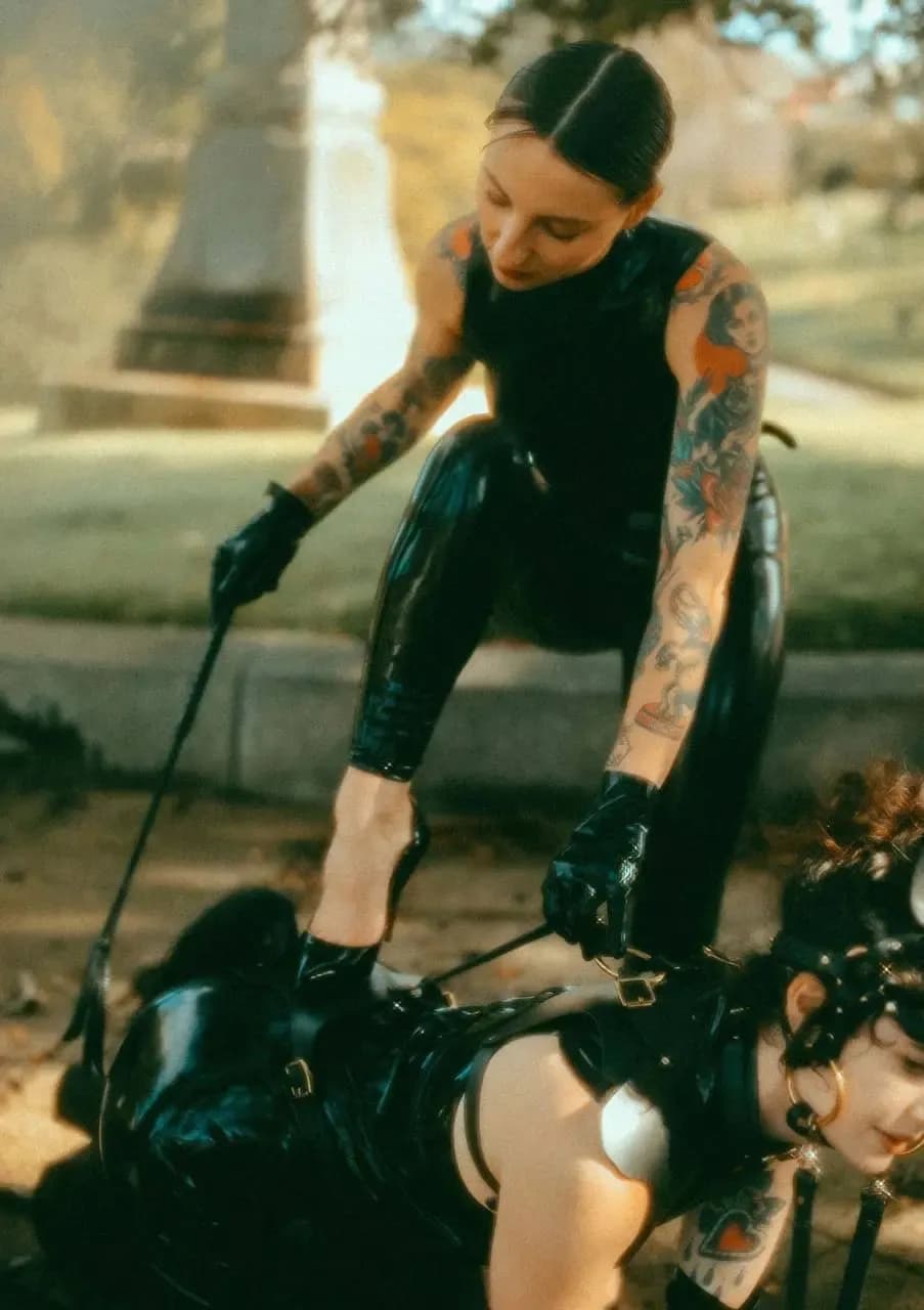 A woman with bangs and red lipstick wearing a black leather jacket with metal studs, revealing floral collarbone tattoos. Behind her stands a person wearing a full black latex hood mask and collar with a chain attached. The foreground is softly blurred.